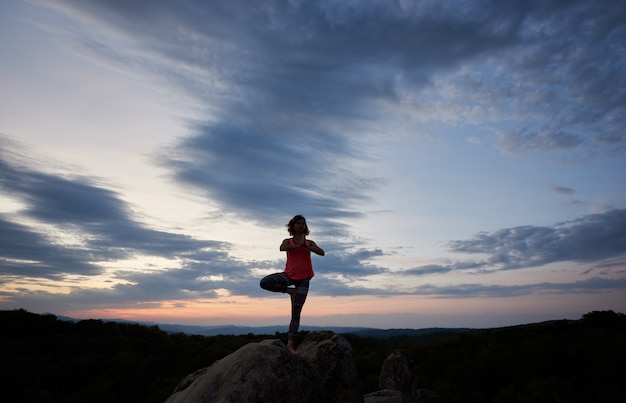 Bild in voller Länge von einer jungen schlanken Frau, die Yogaübungen macht, die barfuß auf einem Bein auf großem Bergfelsen gegen schönen Abendhimmel stehen. Fitness und aktives Lifestyle-Konzept.