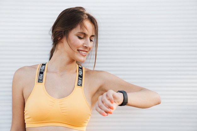 Bild Nahaufnahme einer hübschen Frau in Sportkleidung, die draußen über einer weißen Wand steht und auf die Armbanduhr schaut