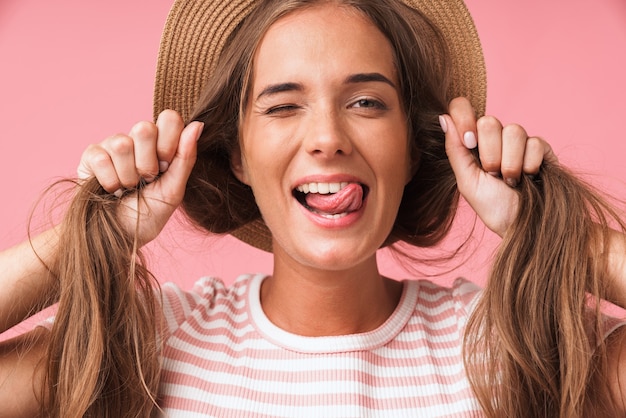 Foto bild nahaufnahme einer jungen fröhlichen frau mit strohhut, die sich mit ihren haaren lustig macht, während sie eine grimasse verzieht und über rosa wand zwinkert?