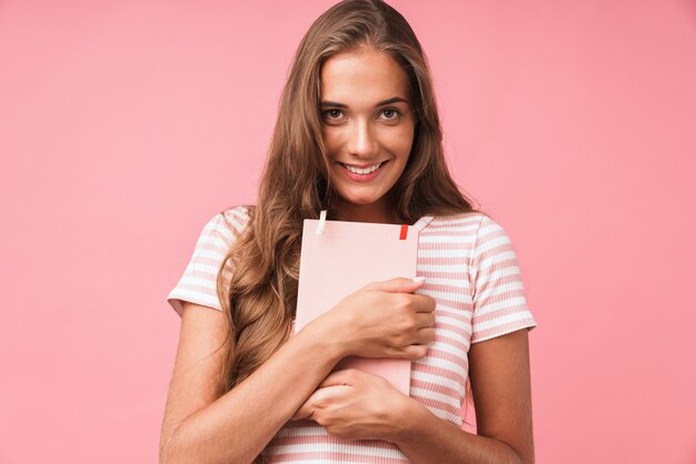 Bild Nahaufnahme eines süßen hübschen Mädchens, das lächelt, während sie ihr Tagebuch umarmt, isoliert über rosa Wand?