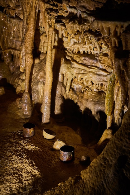 Bild von alten verrosteten Trinkbechern auf dem Boden der Höhle, umgeben von Stalagmiten und Stalaktiten