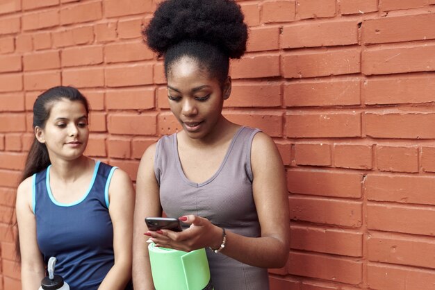 Bild von erstaunlichen jungen athletischen Frauen vor einer Backsteinmauer, die das Telefon untersuchen. Zwei Freundinnen lachen über den Social-Media-Beitrag.