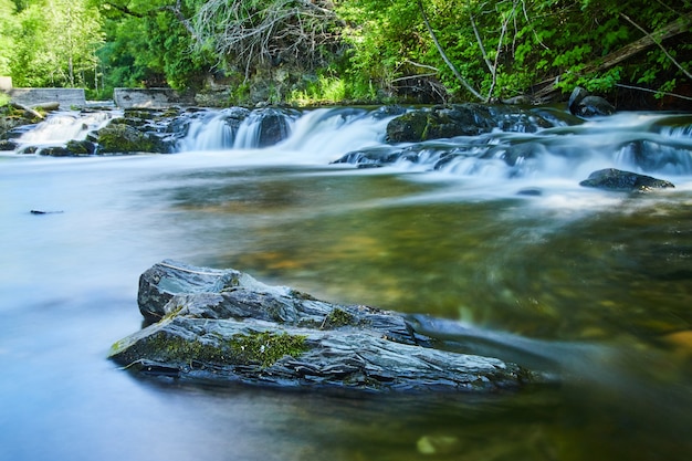 Foto bild von lone rock mit moos auf glattem fluss mit wasserfällen und damm im hintergrund