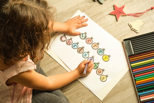 Bild von oben von einem Mädchen, das Frohe Weihnachten-Buchstaben auf weißem Papier ausmalt