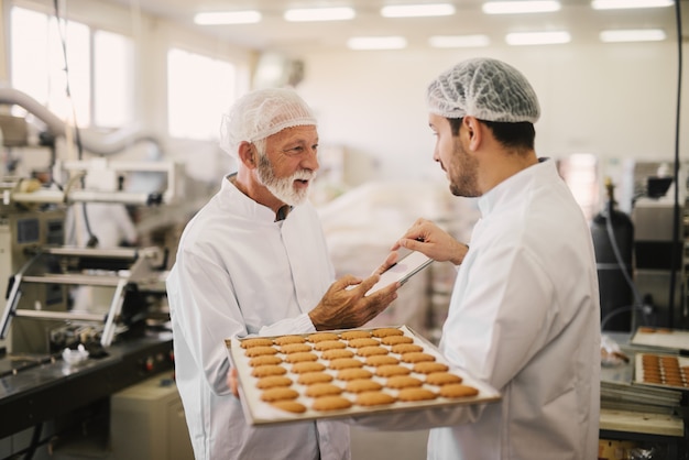 Bild von zwei Mitarbeitern in steriler Kleidung in der Lebensmittelfabrik lächelnd und sprechend. Jüngerer Mann hält Tablett voll mit frischen Keksen, während der ältere Tablette hält und Produktionsprozess überprüft.