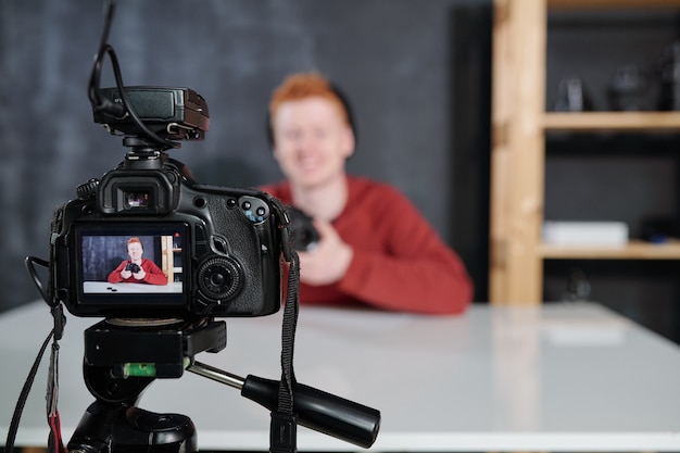 Bildschirm der Videokamera mit dem jungen männlichen Vlogger oder Fotografen, der Fotokamera während des Schießens im Studio hält