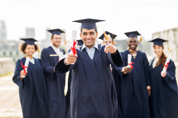 Bildung, Abschluss und Menschenkonzept - eine Gruppe glücklicher internationaler Studenten in Mörtelbrettern und Junggesellenkleidungen mit Diplomen, die mit dem Finger auf Sie zeigen