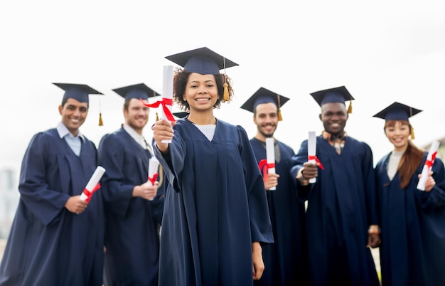 Bildung, Abschluss und Menschenkonzept - Gruppe glücklicher internationaler Studenten in Mörtelbrettern und Junggesellenkleidungen mit Diplomen