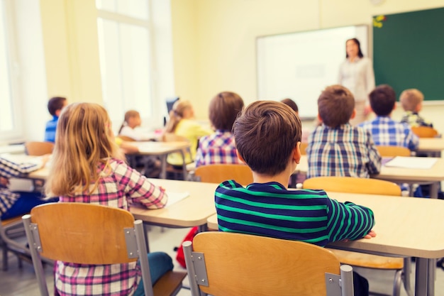 bildung, grundschule, lernen und personenkonzept - gruppe von schulkindern, die sitzen und dem lehrer im klassenzimmer von hinten zuhören