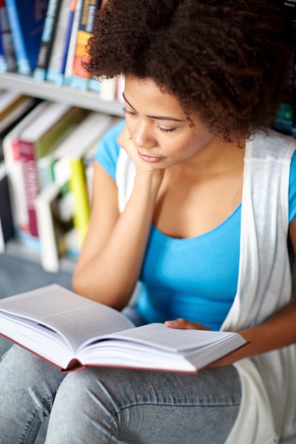 bildung, high school, universität, lernen und personenkonzept - nahaufnahme eines afroamerikanischen studentenmädchens, das buch liest, das auf dem boden in der bibliothek sitzt