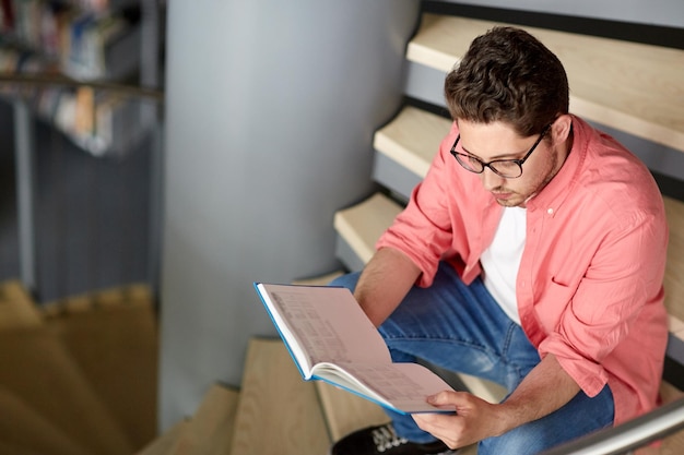 bildung, highschool, universität, lern- und personenkonzept - studentenjunge oder junger mann liest buch, das auf treppen in der bibliothek sitzt