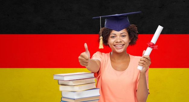 Foto bildung, schule, abschluss, gesten und personenkonzept - glücklich lächelndes afrikanisch-amerikanisches studentenmädchen in bachelor cap mit büchern und diplom, das daumen nach oben über dem hintergrund der deutschen flagge zeigt