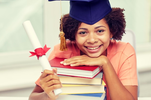 bildung, schule, abschluss und personenkonzept - glücklich lächelndes afroamerikanisches studentenmädchen in bachelor cap mit büchern und diplom, die zu hause am tisch sitzen