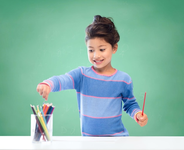 Bildung, Schule, Kinder, Kreativität und fröhliches Menschenkonzept - glückliches kleines Mädchen, das mit Farbstiften auf grünem Kreidetafelhintergrund zeichnet