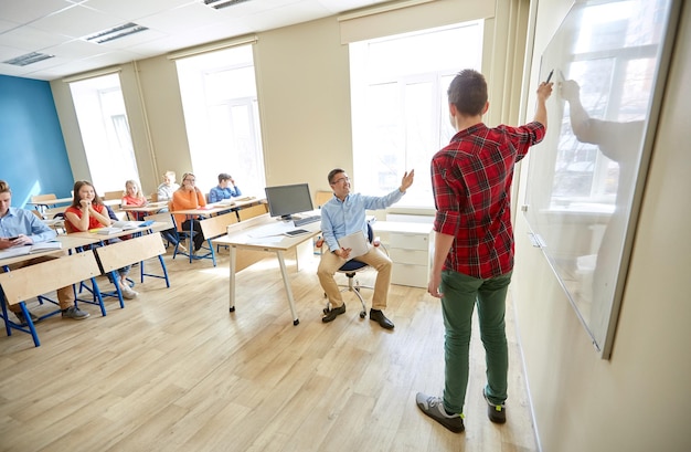 Foto bildung, schule, lernen und personenkonzept - studentenjunge, der etwas auf leerer weißer tafel und lehrer im klassenzimmer zeigt