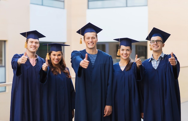 Bildungs-, Abschluss-, Gesten- und Personenkonzept - eine Gruppe lächelnder Studenten in Mortarboards und Kleidern, die im Freien Daumen nach oben zeigen