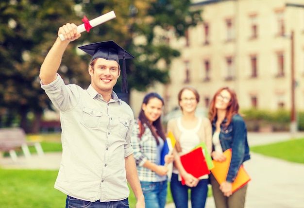 Bildungs-, Campus- und Jugendkonzept - lächelnder Teenager in Eckkappe mit Diplom und Klassenkameraden auf dem Rücken
