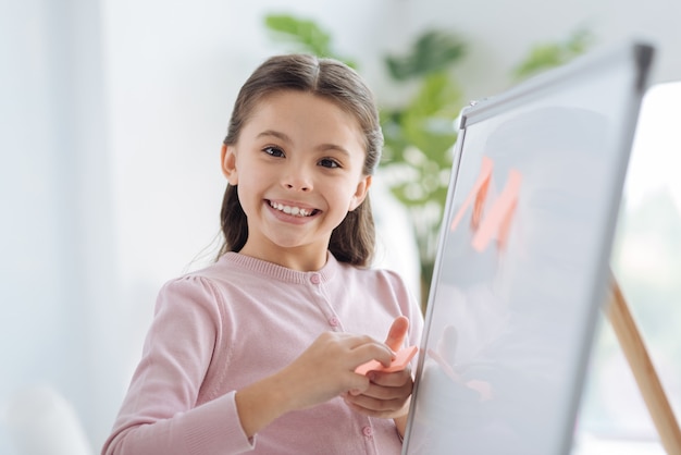 Bildungsprojekt. Nettes positives nettes Mädchen, das nahe dem Whiteboard steht und Haftnotizen darauf setzt, während es dich ansieht