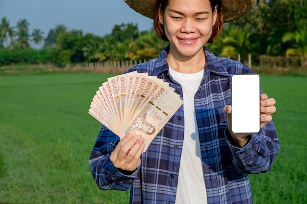 Los billetes tailandeses con un teléfono inteligente de pantalla en blanco son mantenidos por agricultoras en granjas verdes