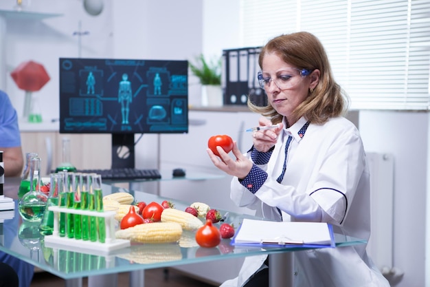 Biólogo de bata blanca en un laboratorio de investigación inyectando líquido en un tomate. Tubos de ensayo con líquido verde.
