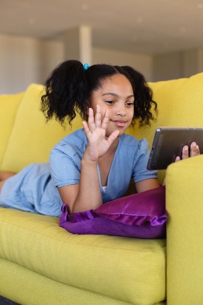 Biracial Grundschülerin winkt über Smartphone während Videogespräch, während sie auf der Couch in der Schule liegt