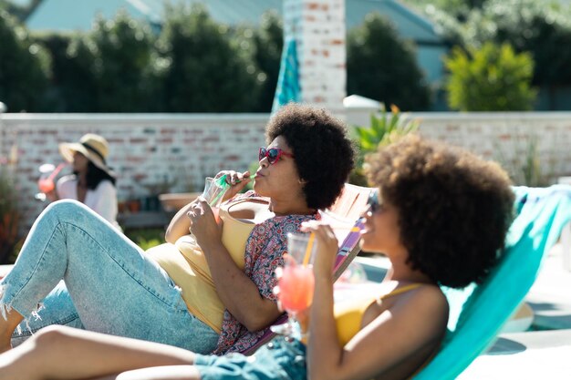 Biracial junge Frauen mit Afro-Haaren trinken Cocktails, während sie auf Liegestühlen bei einer Poolparty entspannen. Treffpunkt, glücklich, unverändert, Freundschaft, zusammen, geselliges Beisammensein, Vergnügen, Wochenendaktivitäten.