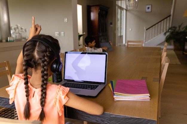 Biracial Mädchen mit Kopfhörern und Laptop mit Kopierplatz für Online-Lektion zu Hause, die Hand hebt