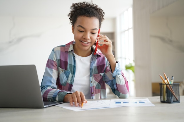 Biracial Teen Girl ruft per Telefon an und macht Hausaufgaben am Schreibtisch mit Laptop Bildung und Technik