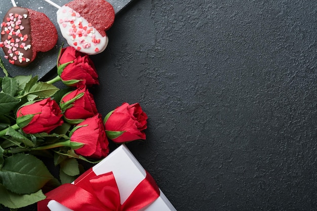 Biscoitos de brownie de flores vermelhas rosas e doces de chocolate na caixa de presente em forma de coração com vinho em fundo preto Dia dos namorados Dia das mães ou cartão de felicitações do Dia das mulheres Vista superior plana leiga com espaço de cópia