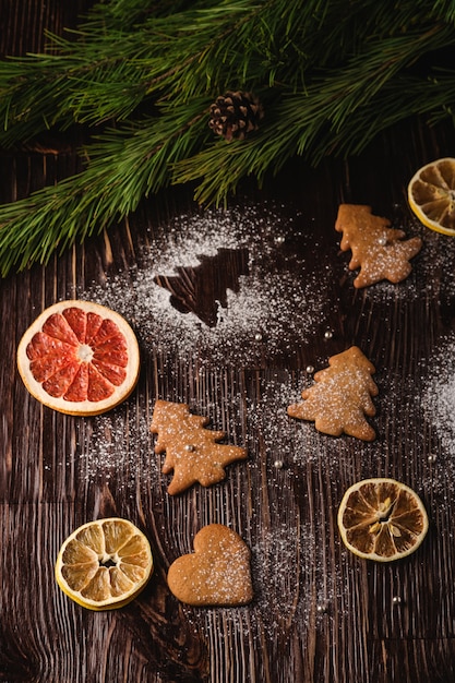 Biscoitos de gengibre em forma de abeto e coração de natal, açúcar em pó na mesa de madeira, frutas cítricas secas, galho de árvore do abeto, vista de ângulo, foco seletivo