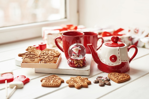 Biscoitos de Natal e caneca de chá quente, tempo de Natal. Pão de Natal, doces, café no copo vermelho na mesa de madeira na mesa de janela de dia gelado de inverno. Férias acolhedoras em casa. Modelo de cartão postal