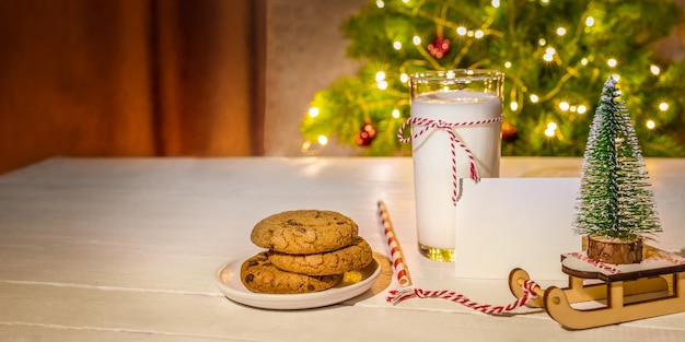 Biscoitos de vidro de leite vazio nota decorações de Natal na mesa branca contra a árvore de Natal com luzes