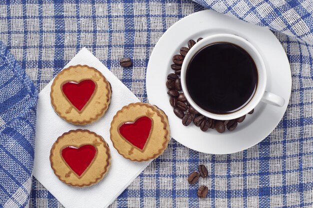 Biscoitos doces com jujuba em forma de coração e uma xícara de café preto na toalha de mesa azul, vista de cima