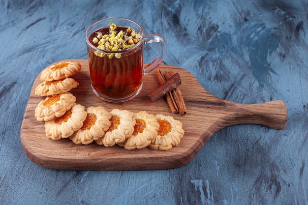 Biscoitos redondos com geleia e um copo de chá preto colocados sobre uma tábua de madeira.