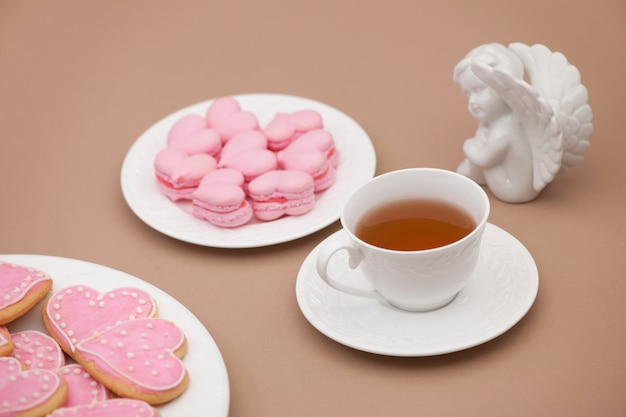 Biscoitos rosa em forma de coração em um prato com uma xícara de chá e um anjo no Dia dos Namorados