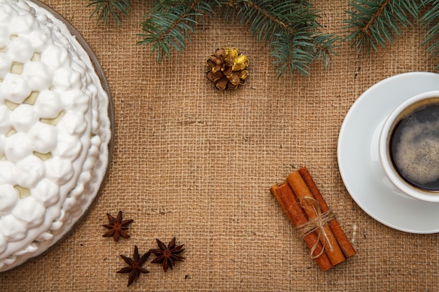 Biskuitkuchen verziert mit Schlagsahne, Tasse Kaffee, Sternanis und Zimt mit Fichtenzweig auf Sackleinen. Ansicht von oben. Frohe Weihnachten-Konzept.