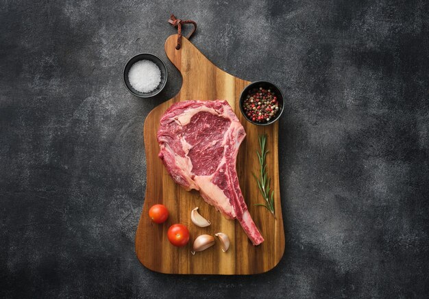 Bistec crudo Tomahawk en tablero de madera con especias para asar fondo gris