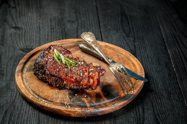 Bistec de Ribeye envejecido seco a la barbacoa con cuchillo y tenedor sobre tabla de cortar. Fondo de madera negra. Naturaleza muerta. Copie el espacio. De cerca