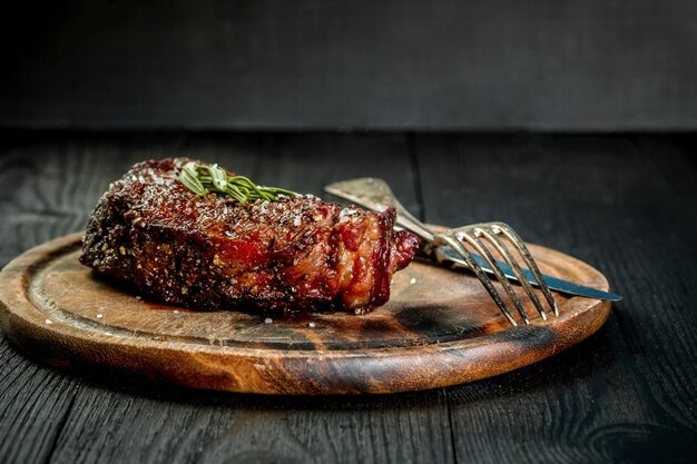 Bistec de Ribeye envejecido seco a la barbacoa con cuchillo y tenedor sobre tabla de cortar. Fondo de madera negra. Naturaleza muerta. Copie el espacio. De cerca