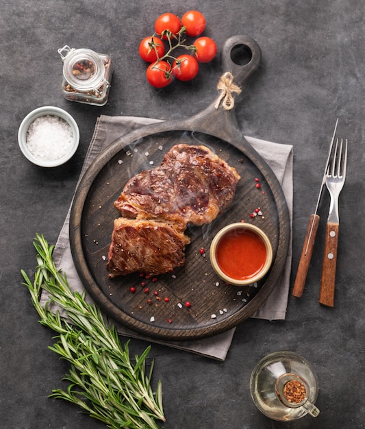 Bistec de ternera asado a la barbacoa con salsa de humo, tomates, sal y romero sobre tabla de madera negra sobre fondo oscuro