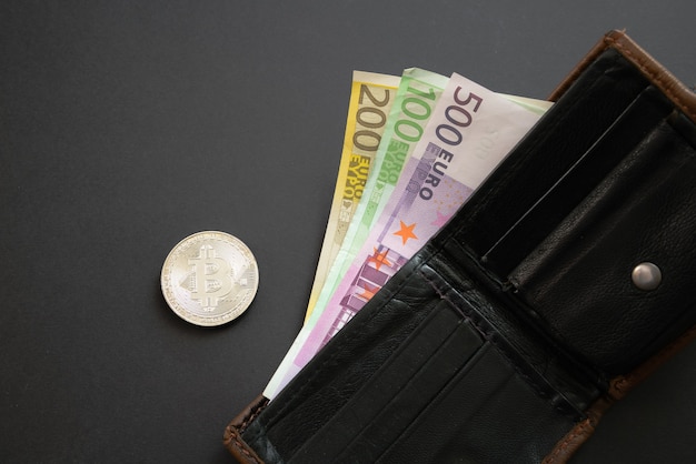 Foto bitcoin neben euro-banknoten, die aus einer brieftasche auf schwarzem hintergrund herausragen. digitale währung, blockchain-markt. euro-scheine neben krypto-münze