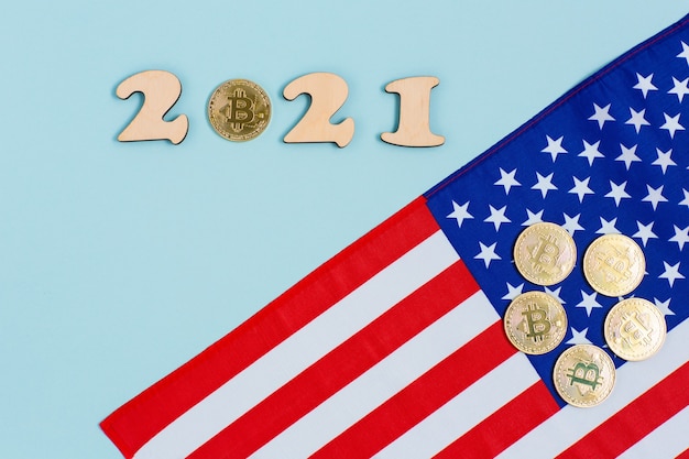 Bitcoins dorados con números de madera 2021 y bandera estadounidense sobre superficie azul