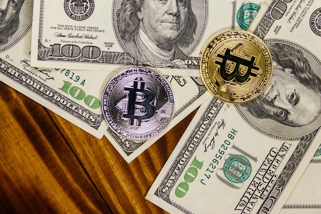 Bitcoins dourados e prateados e notas de cem dólares na mesa de madeira