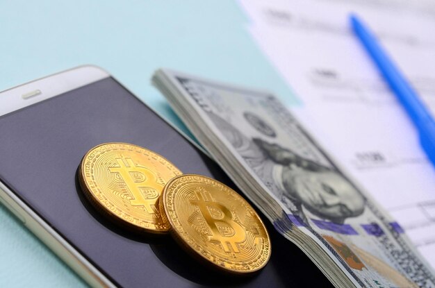 Bitcoins liegt mit den Steuerformularen Hundert-Dollar-Scheine und Smartphone auf hellblauem Hintergrund Einkommensteuererklärung
