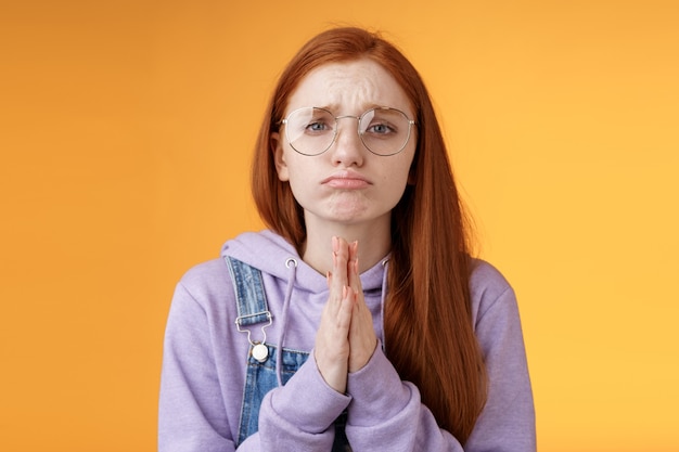 Bitte betteln Sie. Anhänglich verärgert traurige kaukasische rothaarige Freundin mit Brille stirnrunzelnde Grimasse schmollend flehend Hilfe um Gunst bitten dringend etwas ausleihen, orange Hintergrund.