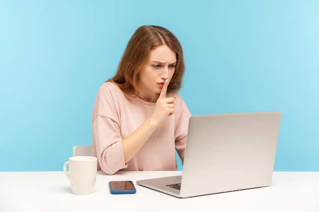 Bitte geheim halten Mitarbeiterin am Arbeitsplatz mit stiller Geste, die über Videoanruf Online-Kommunikation über Laptop im Home Office Indoor Studio Shot isoliert auf blauem Hintergrund spricht