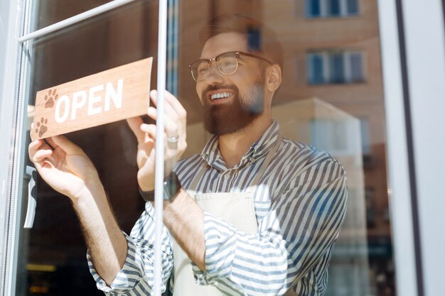 Bitte schön. Fröhlicher, erfreuter Mann, der lächelt, während er das Schild an das Fenster stellt