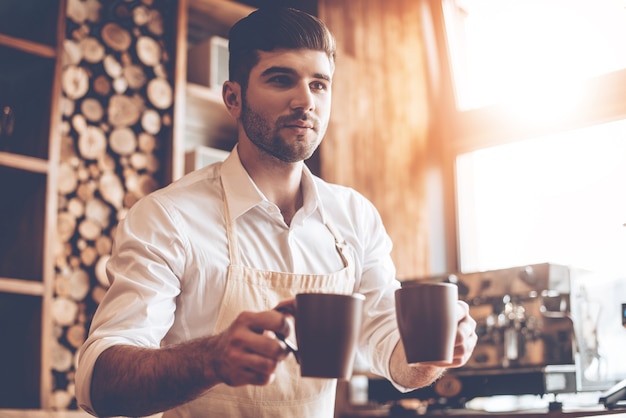 Bitte schön! Junger gutaussehender Mann in Schürze, der zwei Kaffeetassen hält und wegschaut, während er im Café steht