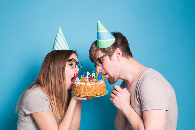 Bizarres junges Paar Mädchen und Junge in Papierhüten wollen ein Stück Glückwunschkuchen abbeißen