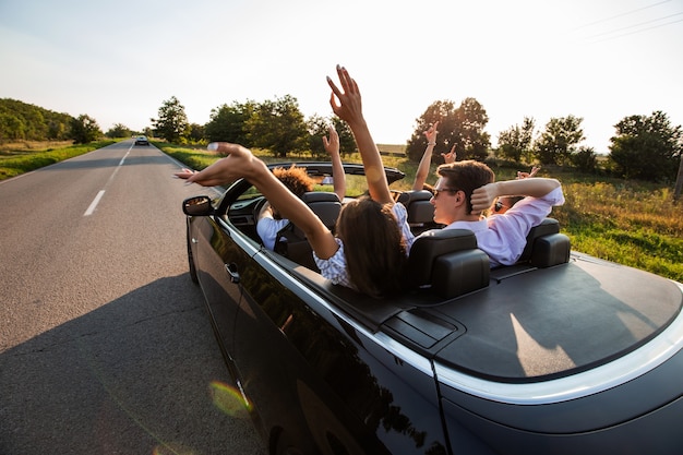 Black cabriolet está na estrada secundária. Companhia de garotas e rapazes estão sentados no carro levantam as mãos em um dia ensolarado. .